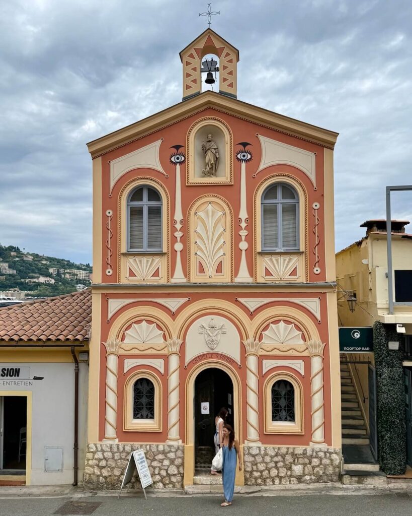 Chapelle Saint-Pierre Cocteau in Villefranche sur Mer - skinny church building from outside in beige and orange with eyes on top and ladies walking out of the door