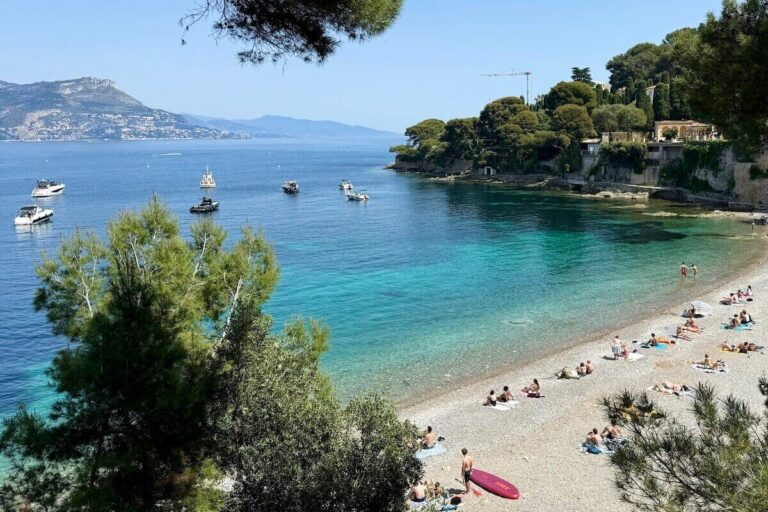 Paloma beach Cap Ferrat on sunny day with green blue water on cove and people lying on towels on the stoney beach - best day trips from nice