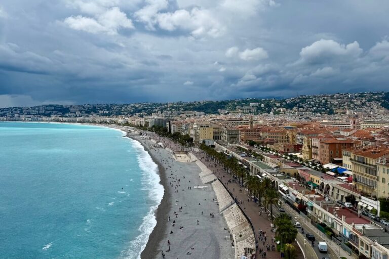 Promenade des Anglais from Castle Hill with turquiose sea, grey stone beach, orange and yellow buildings and cloudy sky - essential stop with one day in Nice