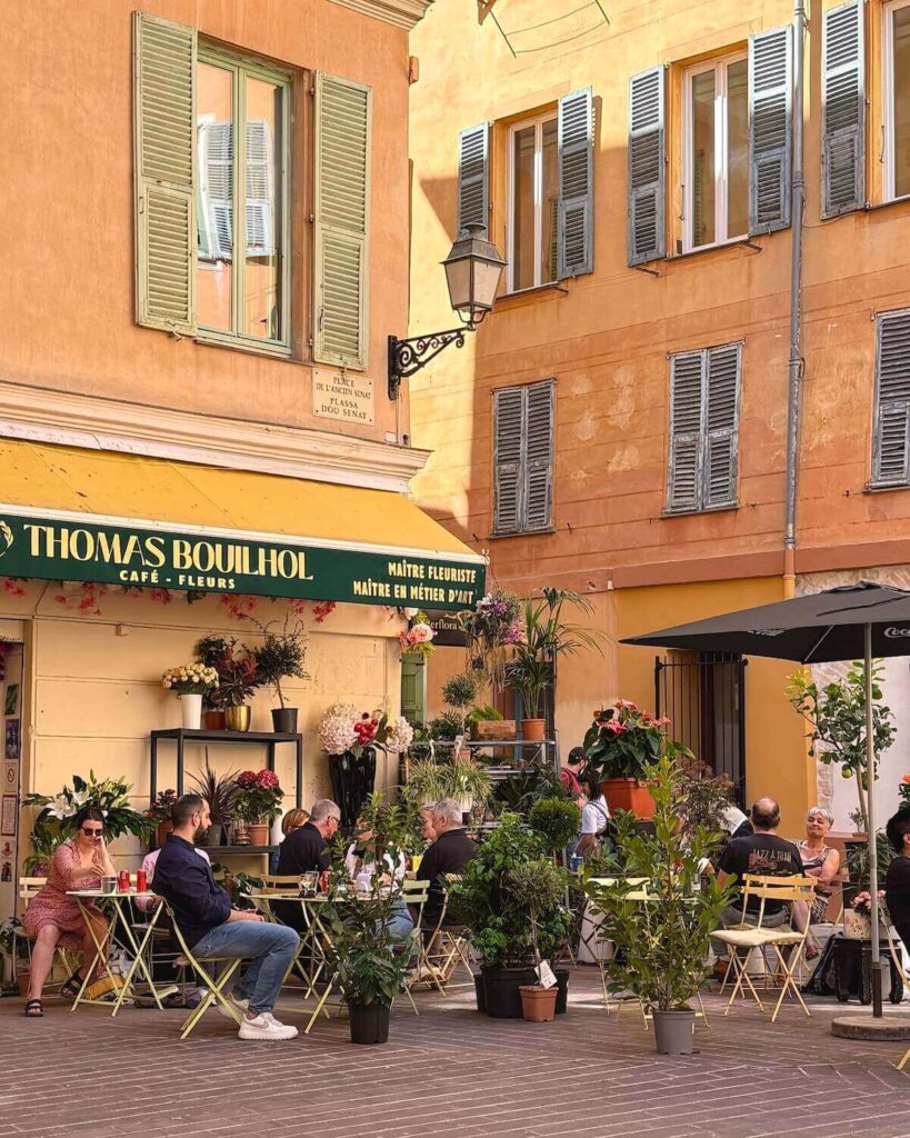 Thomas Boulhol café with patrons at tables and bright coloured buildings and flowers and plants dotted around floor in Nice Old Town