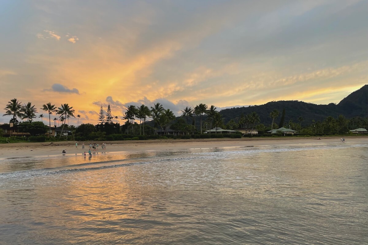 Sunset on Hanalei Bay beach looking from pier towards beach with palm trees and mountainous background - one of the best things to do in Kauai